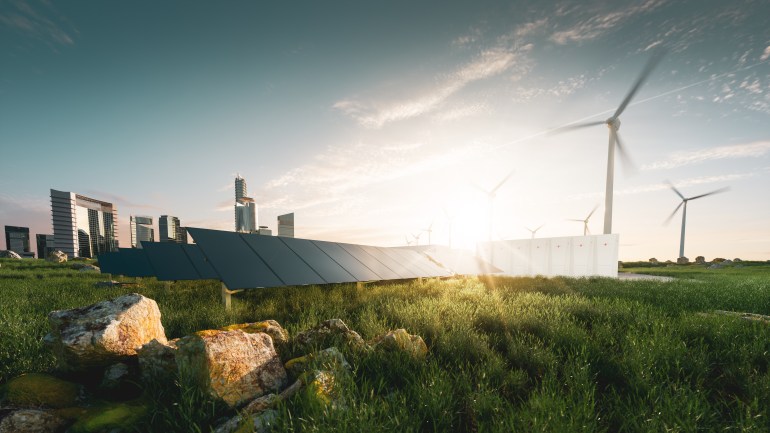 Concept of sustainable energy solution in beautifull sunset backlight. Frameless solar panels, battery energy storage facility, wind turbines and big city with skycrapers in background.