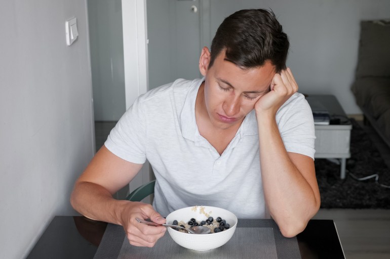 sleepy sunburned man in t-shirt sits at table resting head with hand and eats healthy breakfast at hotel restaurant in morning; Shutterstock ID 1714895911; purchase_order: aljazeera ; job: ; client: ; other: