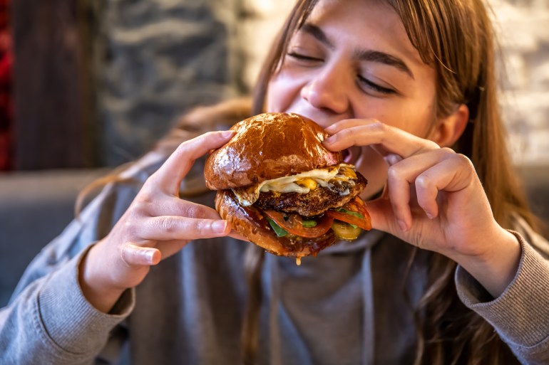 Close-up of a girl eating a big burger with meat and vegetables on a blurred background.