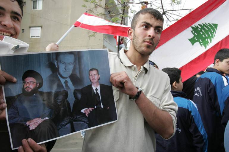 Standing in front of Lebanese national flags, two young men carry a picture of Syrian President Bashar al-Assad (R) and the secretary general of the Lebanese Shiite Muslim group Hezbollah, Sheikh Hassan Nasrallah with a portrait of Syria's late leader Hafez al-Assad in the background, during an anti-US protest outside the United States' embassy in the Beirut northeastern suburb of Awkar, 30 March 2005. Washington said yesterday it opposed any suggestion that Lebanese parliamentary elections scheduled for May be postponed because of continuing political turmoil in the Middle East country. AFP PHOTO/JOSEPH BARRAK (Photo by Joseph BARRAK / AFP)