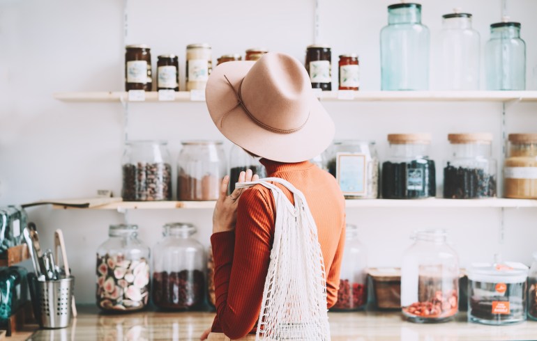 Minimalist vegan style young woman with cotton reusable net mesh bag on background of goods of zero waste shop. Shopping without plastic packaging in plastic free grocery store.