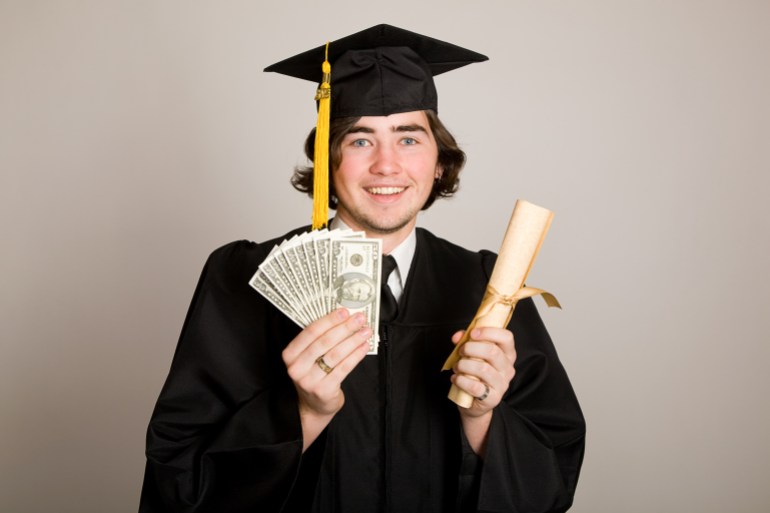 A young man in a graduation cap & gown holding a handful of money and a diploma.